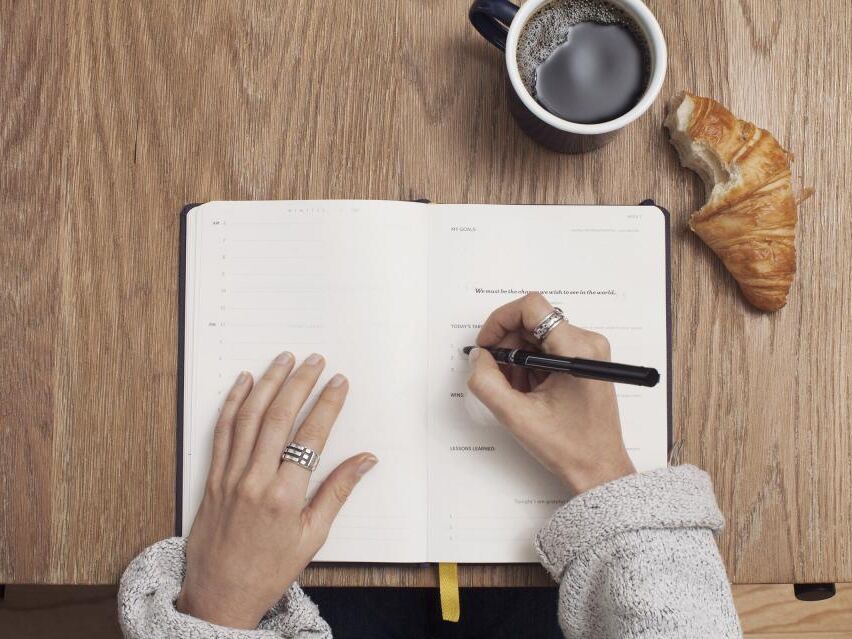 Mãos a escrever numa agenda aberta, ao lado de um café e de um croissant meio comido